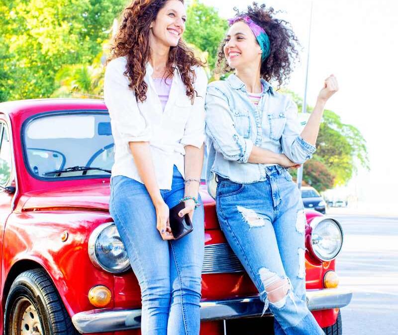 Two cheerful women with curly hair lean against the hood of a vintage red car, smiling warmly at each other - www.beatriceinsuranceagent.com